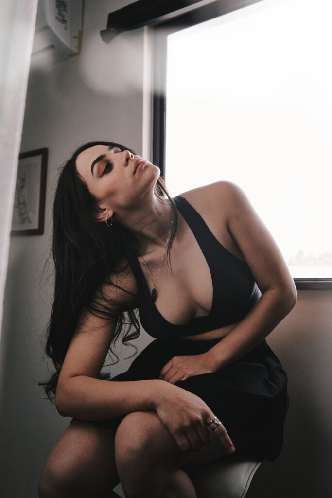 Valeria Cisneros poses elegantly by a window wearing a black top and skirt with a necklace and rings bathed in soft light.